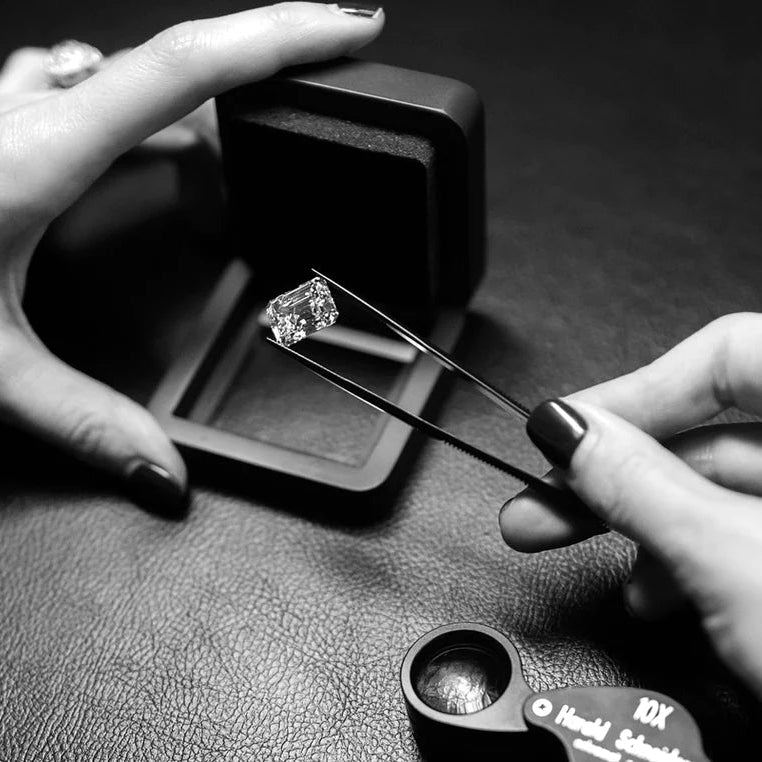 A hand holds tweezers with a loose lab-grown diamond above an open jewelry box, with a jeweler's loupe on a textured surface.